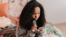 Young woman wearing a sweatshirt is sitting on her bed and checking a mobile application for information.