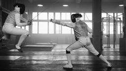 Black and white image of two individuals fencing. One is jumping but the opponent finds the weak spot. 