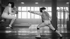 Black and white image of two individuals fencing. One is jumping but the opponent finds the weak spot. 